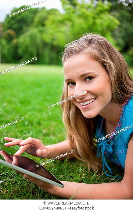 Smiling teenager lying down on the grass in a parkland while touching her tablet pc