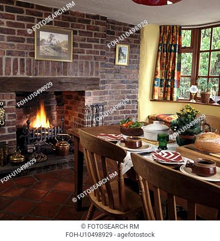 Lighted fire in fireplace in exposed brick wall in cottage dining room with table set for lunch