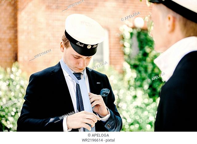 Male university student adjusting necktie while standing with friend at graduation party