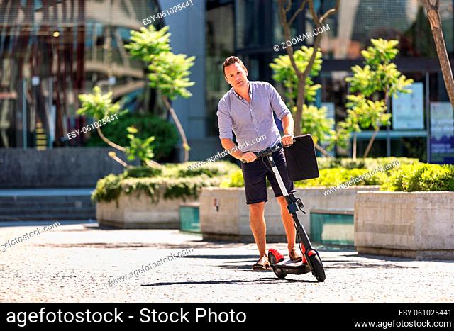 Young casual active sporty businessman in urban city center, wearing shirt and shorts, holding laptop bag, riding to work on electric scooter on a hot summer...