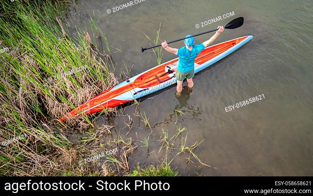 senior male paddler is stretching before workout on his racing stand up paddleboard, aerial view of a lake shore