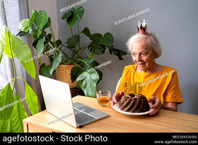 Happy senior woman showing birthday cake on video call through laptop at home
