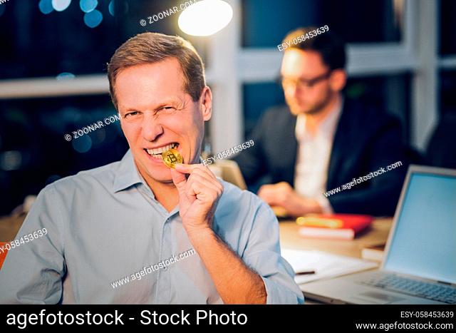 Funny mature man bites shiny golden bitcoin. Middle aged man wearing blue coloured shirt angrily biting golden bitcoin coin with his coworker working in...