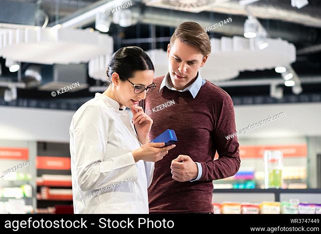 Experienced female pharmacist checking the indications and contraindications of a new medicine next to a young male customer in a modern pharmacy