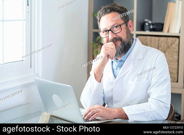 Smiling old male doctor in glasses and white uniform sit at desk in hospital work on laptop write in journal, happy mature senior man physician fill patient...