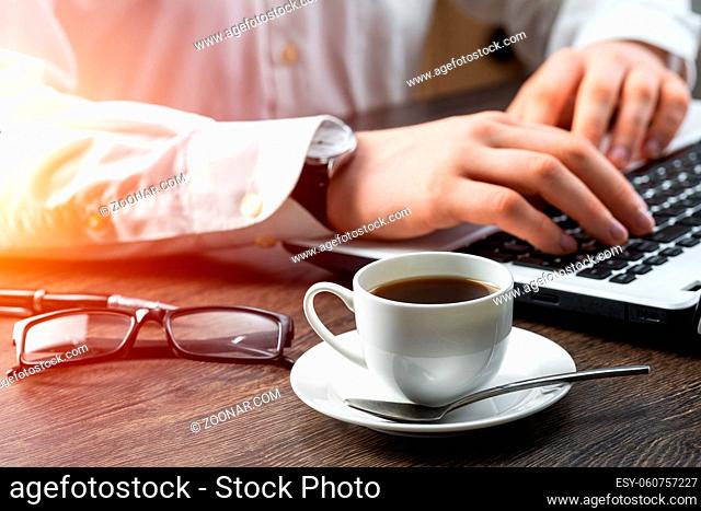 Businessman sitting at desk and using laptop. Close-up of man hands typing on keyboard in office. Side view consultation workplace with glasses and coffee cup