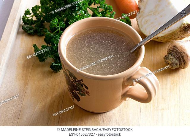 Chicken stock in a vintage mug with a spoon, with parsley, carrot and celery root in the background