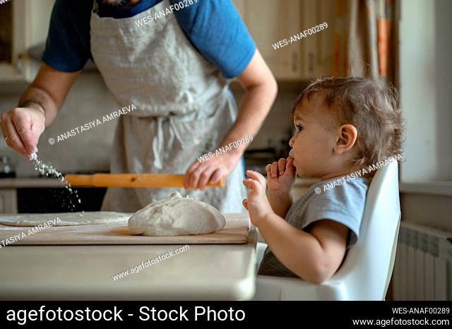 Son sitting by father rolling pizza dough in kitchen