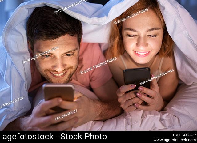 happy couple using smartphones in bed at night