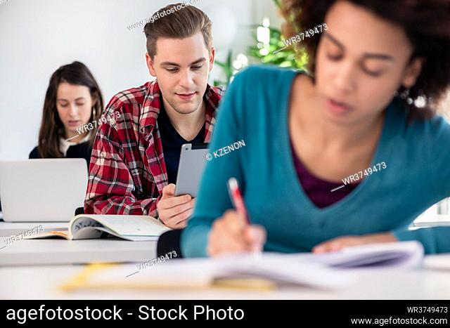 Young handsome student smiling with a nostalgic facial expression while using a tablet during class in a modern college or university
