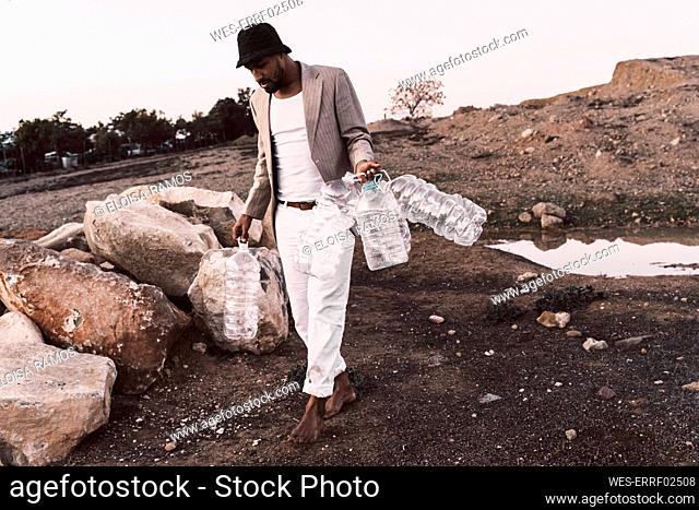 Young man walking in barren land holding empty plastic bottles