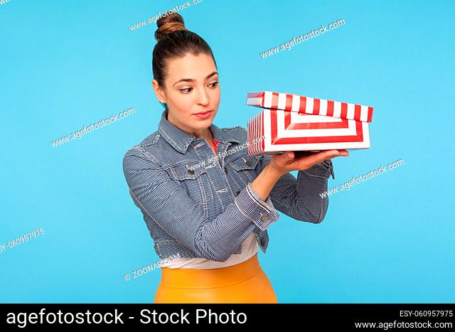 Positive curious nosy woman with hair bun in denim jacket unpacking and opening small carton box, looking inside with interest, hoping for present
