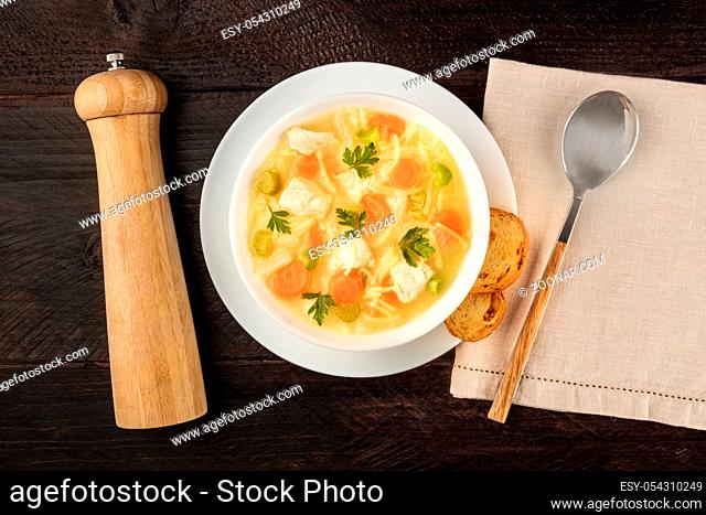 Chicken noodle soup, shot from above on a dark rustic wooden background with toasted bread and a pepper mill
