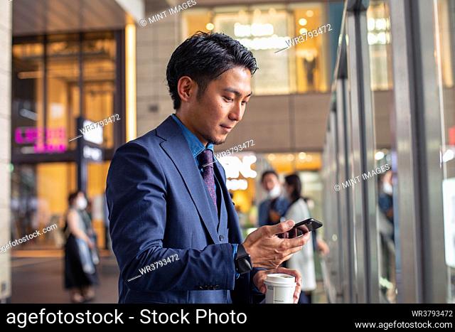 A young businessman in a blue suit in a city, looking at his mobile phone screen, texting or reading a message