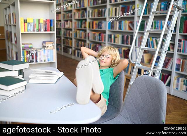 Calm relaxed cute blonde teenage boy sitting on the chair with his feet put up on the library table