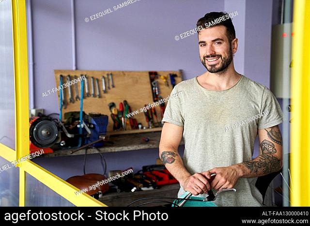 Medium-shot of smiling bearded mechanic standing in his workshop in front of his tools
