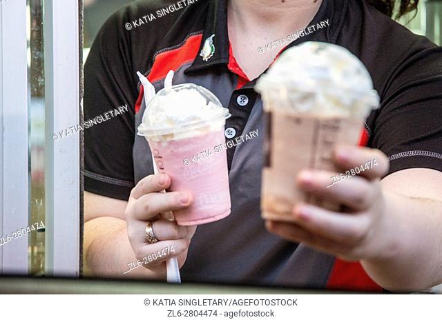 A caucasian fast food employee hands through the drive in a chocolate shake/icecream and a strawberry ice-cream with straws to the consumer in the car