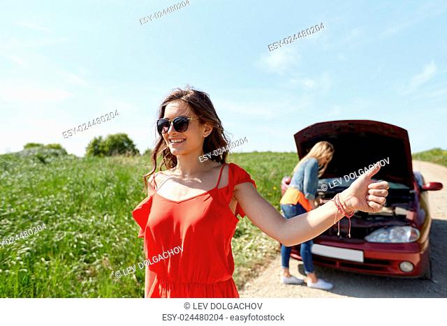 road trip, transport, travel, gesture and people concept - young women with broken car showing hitchhiking gesture asking for help at countryside