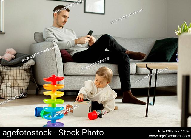 Toddler girl playing in living room, father sitting on sofa