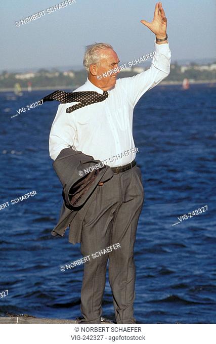 full-figure, white haired senior, ca. 70 years old, wearing a white shirt and tie stands with his jacket hanging over his arm waving near the water  - GERMANY