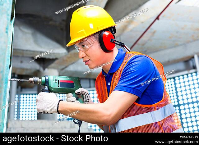 Asian Indonesian builder or worker drilling with a machine or drill, ear protection and hardhat or helmet in a wall of a tower building or construction site