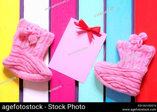 Congratulations on childbirth concept with a message card tied with a red bow, surrounded by cute knitted bootees, on a colorful wooden background