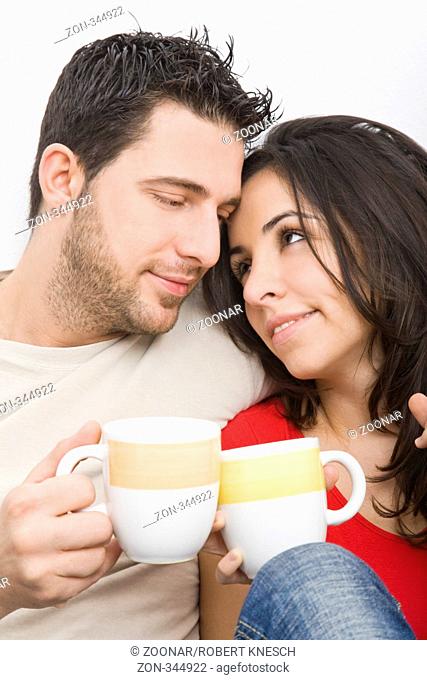 Junges Paar sitzt aneinandergekuschelt an einer Wand gelehnt und hält zwei Teetassen in der Hand Model l.: Kosta Lales