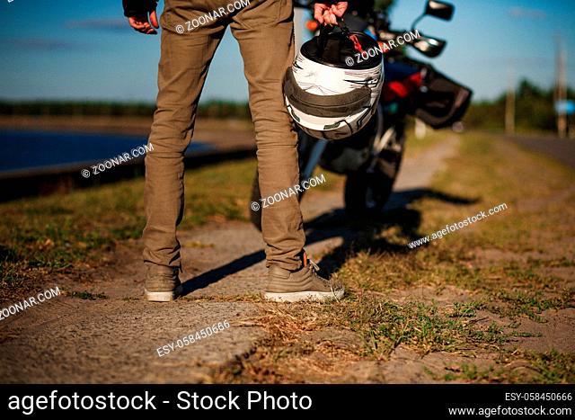 A biker stands near his motorcycle on an empty road. Close view on motorcycler's legs and helmet