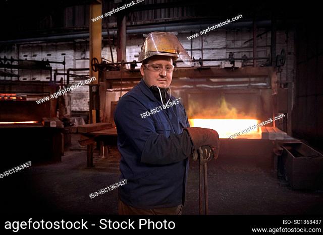 Forge worker, second in command of hammer control or stamper in pre forming forging area with a set of industrial tongs as furnace door is closed