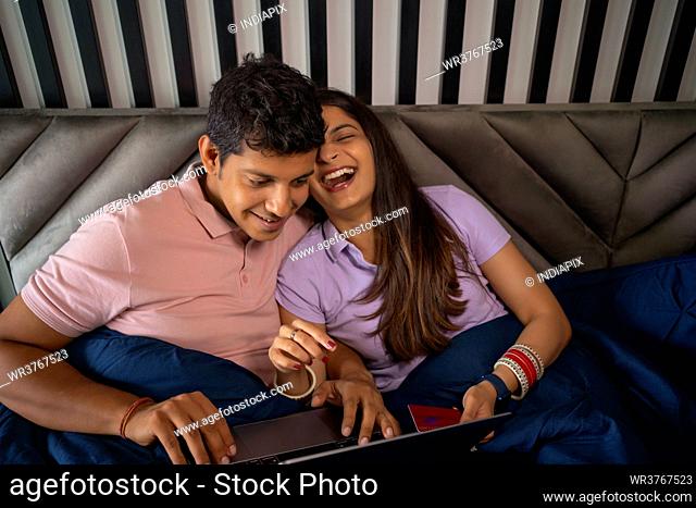 Young newlyweds laughing while shopping online with credit card and laptop