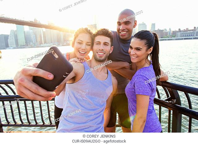 Four adult running friends taking smartphone selfie on riverside, New York, USA