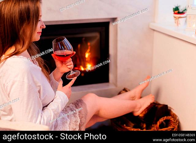 Side view of a young brunette girl is sitting by the fireplace. The beautiful girl is sitting in a cozy room with a glass of red wine