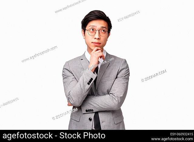 Waist-up portrait of smart and creative young asian male entrepreneur in grey suit, making plan, hold hand on chin and looking up thoughtful