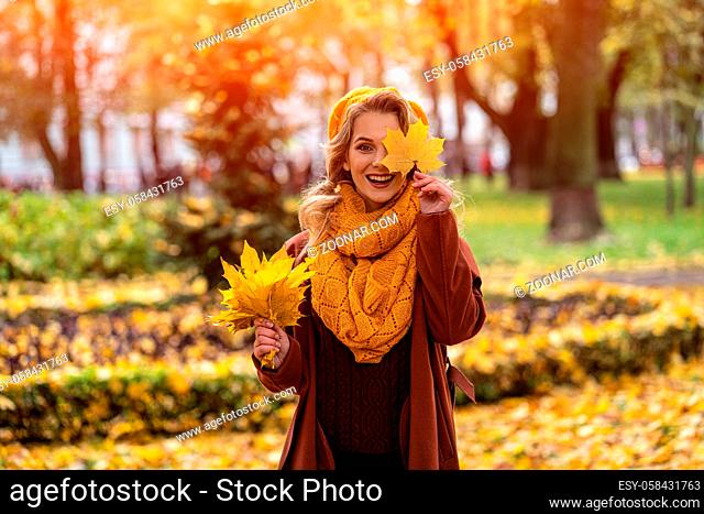 Happy hiding eye woman with a yellowed leaf in yellow knitted beret with autumn leaves in hand and fall yellow garden or park