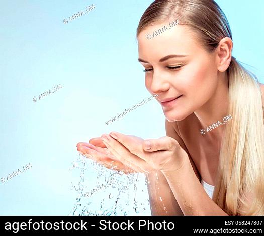 Portrait of a young beautiful woman cleaning her face with fresh cold water, isolated on blue background, morning freshness, removing makeup