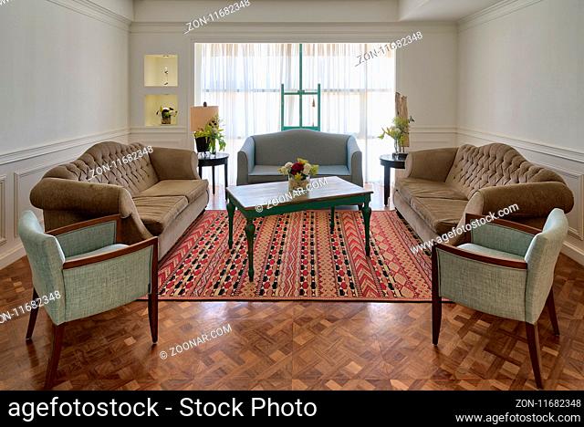 Interior shot of luxury modern living room with three couches, two armchairs, and vintage wooden table on background of big window with white curtains and...