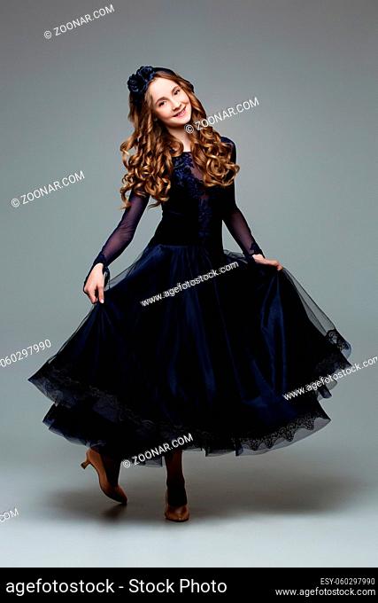Beautiful teenager ballroom dancer with long blond hair in long dark blue dress making curtsy. Studio shot on grey background. Copy space