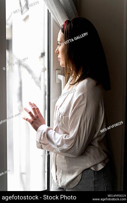 Thoughtful young woman looking though window while standing at home
