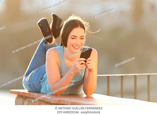 Full body portrait of a happy woman using a smart phone lying in a balcony