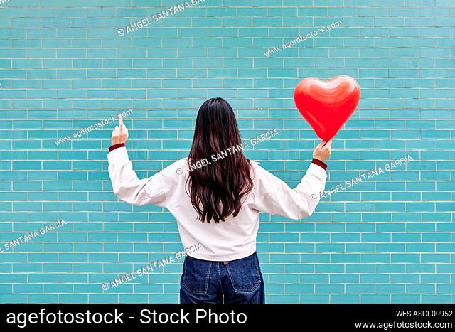 Young woman showing middle finger while holding red balloon in front of brick wall