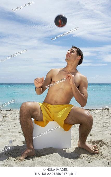Young man sitting on an ice box on the beach and playing with a soccer ball