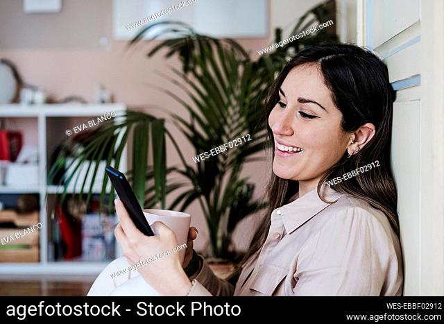 Smiling mid adult woman using smart phone while holding coffee cup at home