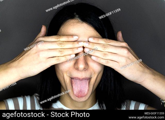 Young woman covering eyes with hands while sticking out tongue against gray background