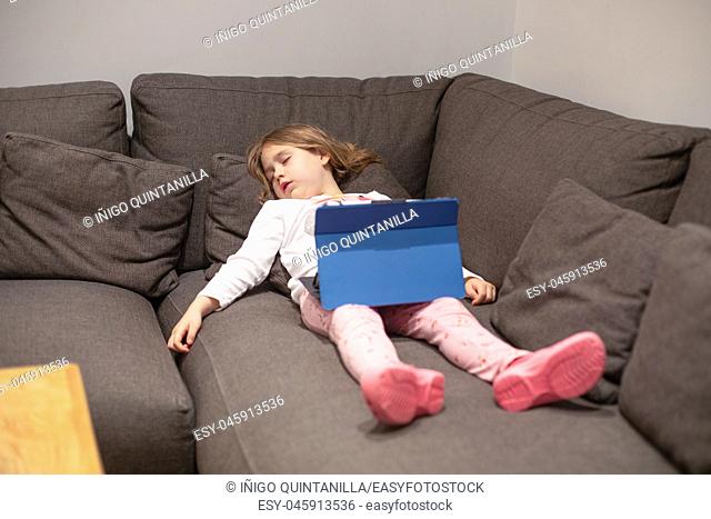 five years old blonde girl sleeping lying on the coach in living room of home, watching digital tablet on her legs