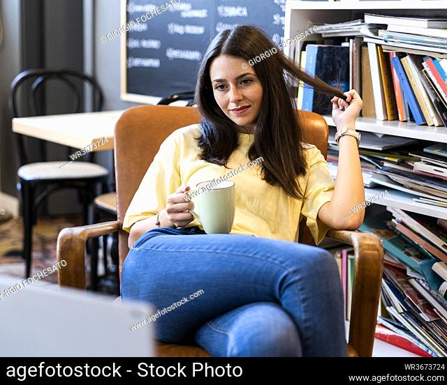 Beautiful young woman holding coffee mug while sitting on armchair in coffee shop