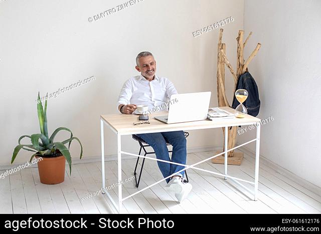 Good mood. Gray-haired man with coffee smiling at screen of laptop sitting at table in modern bright room