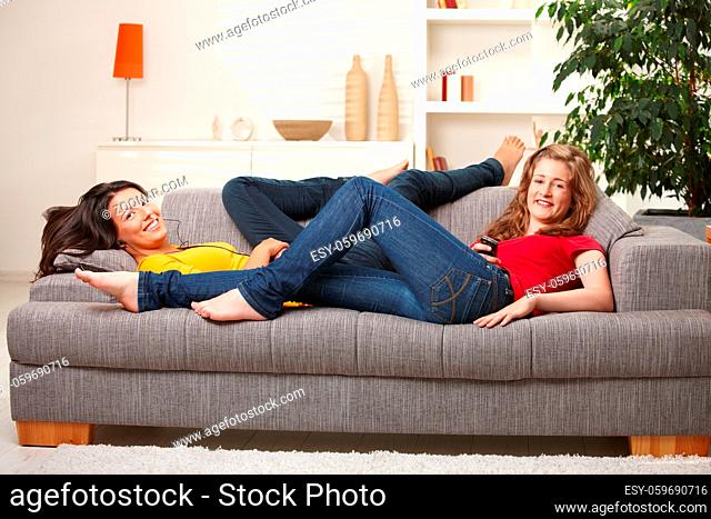 Happy teen girls lying on sofa together smiling at camera having rest at home