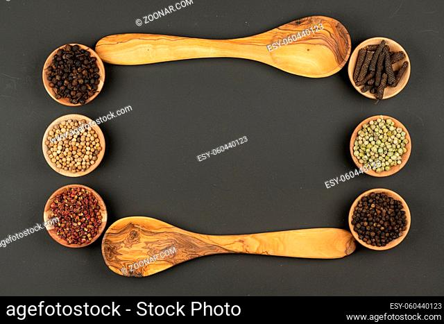 Six small wooden bowls filled with various types of peppercorns and two wooden cooking spoons made of olive wood lie on a black background with copy space in...