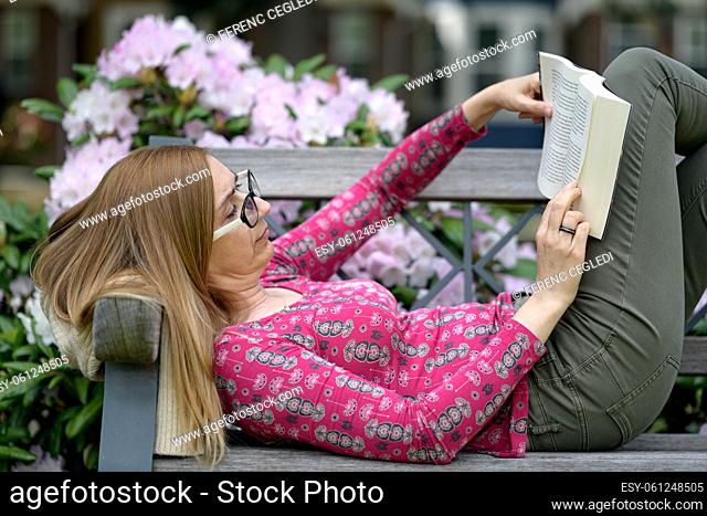 Garden city woman enjoying the sunshine and fresh air filled with spring flower scent while lying on a bench and reading a book outside where the streets are...