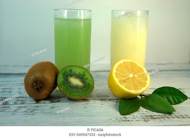 Two glass glasses with a refreshing juice and ice, on a textile stand, whole and sliced half of an orange with leaves and kiwi, lies on a white wooden table